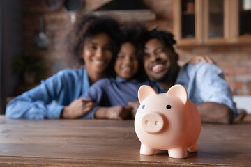 Close up piggybank standing on table with blurred happy African American couple parents and small...