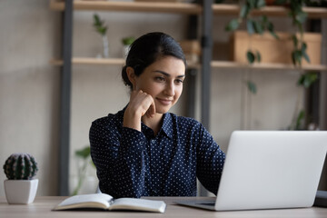 Happy pensive young Indian ethnicity businesswoman looking at computer screen, working on online...
