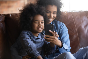 Happy bonding young African American mother and little cute multiracial child daughter using cellphone, playing mobile games, choosing goods shopping in internet store, resting together at home.