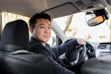 A man driving an Asian car behind the wheel looks at the camera and smiles