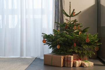 Fototapeta na wymiar Small christmas tree with gifts in paper wrap near window