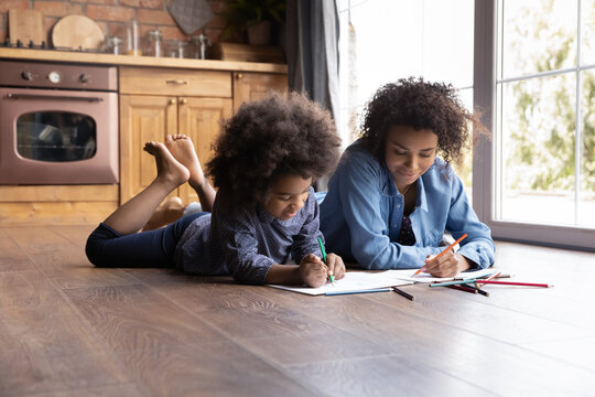 Happy bonding family little African ethnicity child girl involved in creative activity with happy young biracial mother, drawing pictures in paper album lying together on heated floor.