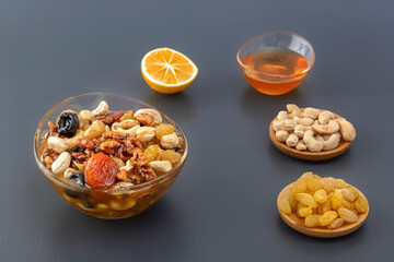 Mixture of walnuts, cashews, raisins, dried apricots and prunes with honey in a glass jar with ingredients and lemon on a gray table