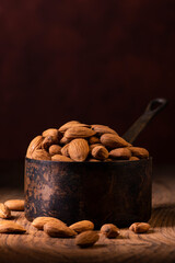 In the foreground, a group of shelled almonds in an aged metal measuring cup against a dark background