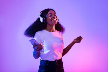 Happy African American woman with headphones and smartphone listening to music and dancing in neon...