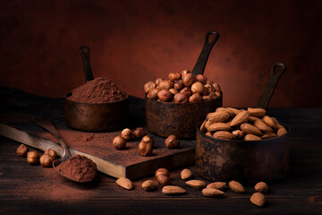 Still life. in the foreground with dark background, vintage metal measuring spoons with shelled hazelnuts and almonds and cocoa powder - 470433162