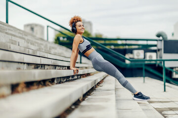 A cheerful sportswoman is listening to music while doing a reverse plank on the stairs downtown. An urban sportswoman exercising outdoors.