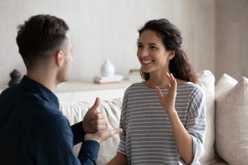 Smiling millennial Latina woman communicates with man use sign language sit together on sofa at home. Couple or friends with disability talking, showing fingers gestures share news, enjoy conversation