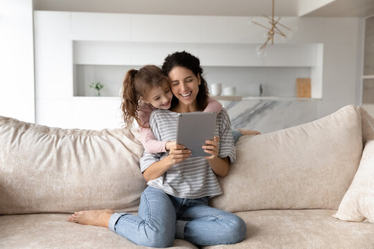Smiling mother with cute daughter having fun with tablet together, looking at screen, chatting online by video call or watching cartoons, young mom piggy backing adorable girl kid, enjoying weekend