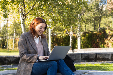 Beautiful business woman with glasses smiling while working on a laptop while sitting in the park. Young professional redhead woman outside the office. Copy space for text