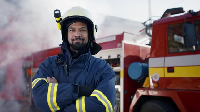 Happy young American-African firefighter man looking at camera with fire truck and smoke in background