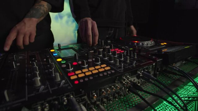 Close-up of multiethnic djs hands controlling sound mixer and cd player turntables with illuminated buttons, mixing musical tracks while performing live techno dance music at stage in nightclub.