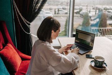 a brunette business woman is sitting at a table in a cafe with a laptop and shopping online with a...