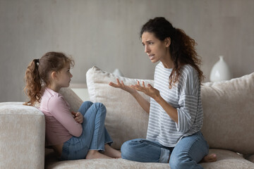 Annoyed emotional strict mother lecturing offended little daughter for bad behavior, sitting on couch at home, unhappy serious mom scolding shouting upset child, family generation conflict concept - Powered by Adobe