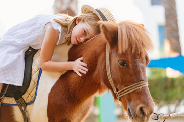 a little girl in a white dress and straw hat sits on top of a pony and hugs.