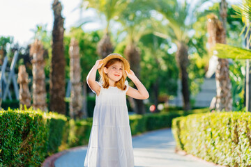 Naklejka premium little girl in a white dress and a straw hat on the path in a tropical hotel