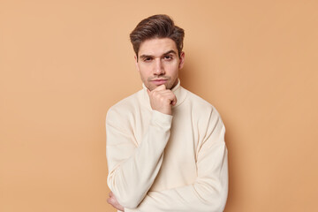 Serious focused man holds chin listens carefully information or curious plan holds chin thinks about something stands determined and assertive wears casual jumper isolated over beige background.