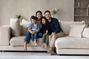 Beautiful young 35s couple and little children sit on cozy couch in living room smiling looking at camera pose for family portrait. Happy homeowners and bank loan, upbringing and kids adoption concept