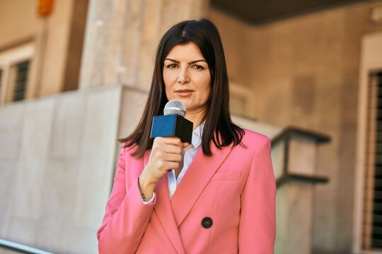 Middle age reporter woman holding microphone doing television speech outdoors