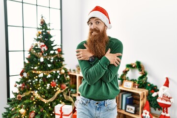 Redhead man with long beard wearing christmas hat by christmas tree shaking and freezing for winter cold with sad and shock expression on face