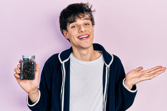 Handsome hipster young man holding jar with coffee beans celebrating achievement with happy smile and winner expression with raised hand