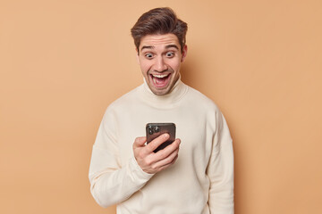 Positive brunet adult man looks happily at smartphone screen reads amazing notification wears...