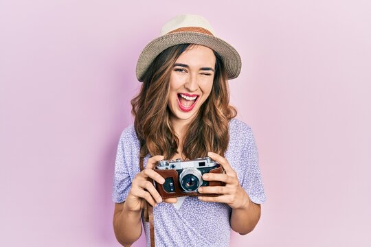 Young hispanic girl wearing summer hat holding vintage camera winking looking at the camera with sexy expression, cheerful and happy face.
