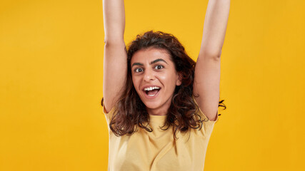 Portrait of excited young woman with dark curly hair in casual wear looking cheerful at camera posing with arms raised isolated over yellow background