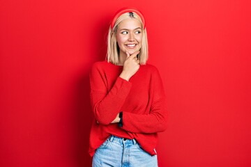 Beautiful blonde woman wearing casual red sweater with hand on chin thinking about question, pensive expression. smiling with thoughtful face. doubt concept.