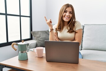 Young brunette woman using laptop at home drinking a cup of coffee with a big smile on face, pointing with hand and finger to the side looking at the camera.