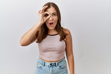 Obraz na płótnie Canvas Young brunette woman standing over isolated background doing ok gesture shocked with surprised face, eye looking through fingers. unbelieving expression.