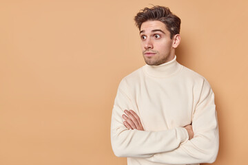 Studio shot of handsome man with dark hair keeps arms crossed looks surprisingly away reacts on shocking scene wears casual jumper isolated over beige studio wall with blank copy space away.