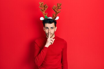 Young hispanic man wearing cute christmas reindeer horns asking to be quiet with finger on lips....