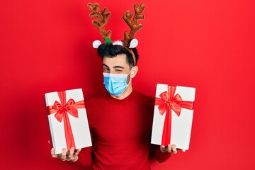 Young hispanic man wearing cute christmas reindeer horns and medical mask holding gifts winking...