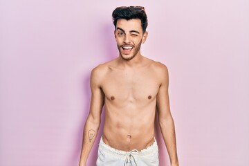 Fototapeta premium Young hispanic man wearing swimwear shirtless winking looking at the camera with sexy expression, cheerful and happy face.