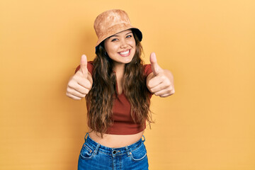 Obraz na płótnie Canvas Young hispanic girl wearing casual clothes and hat approving doing positive gesture with hand, thumbs up smiling and happy for success. winner gesture.
