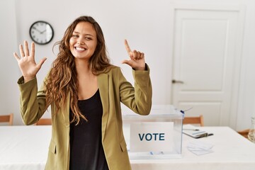 Beautiful hispanic woman standing at political campaign room showing and pointing up with fingers number seven while smiling confident and happy.