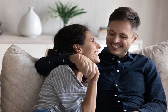 Young 30s peaceful couple hugging relaxing on sofa, enjoy conversation laughing feel carefree spend romantic date indoors. Happy homeowners family leisure at modern home, tenant and bank loan concept