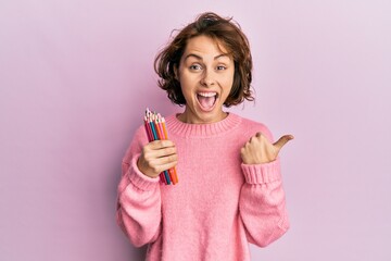 Young brunette woman holding colored pencils pointing thumb up to the side smiling happy with open...