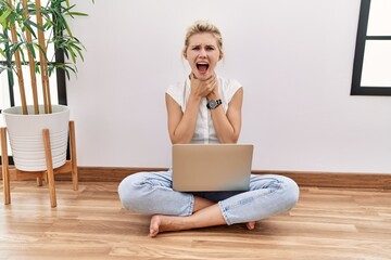 Young blonde woman using computer laptop sitting on the floor at the living room shouting and...