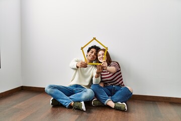 Young hispanic couple holding house project sitting on the floor at empty new home.