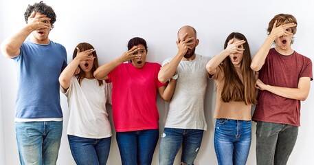 Group of young friends standing together over isolated background peeking in shock covering face and eyes with hand, looking through fingers with embarrassed expression.