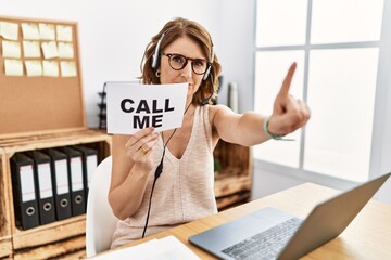 Middle age brunette woman wearing operator headset holding call me banner pointing with finger up...