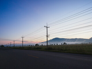 Fototapeta na wymiar Country road at sunrise and electric wires with blue sky, mountains in the background