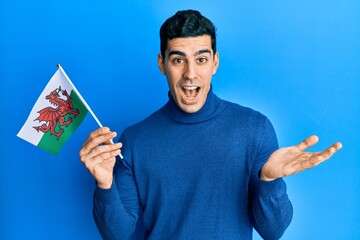 Handsome hispanic man holding wales flag celebrating achievement with happy smile and winner expression with raised hand