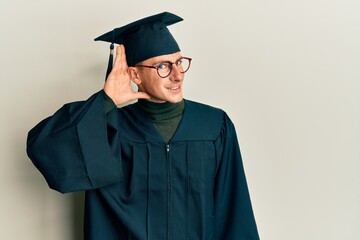 Young caucasian man wearing graduation cap and ceremony robe smiling with hand over ear listening an hearing to rumor or gossip. deafness concept.