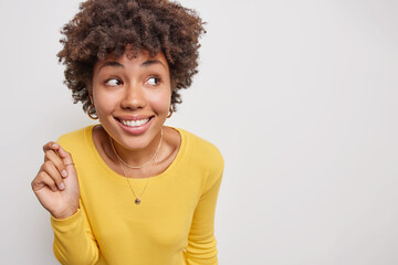 Cheerful young woman with dark curly hair smiles gladfully concentrated away wears yellow jumper...