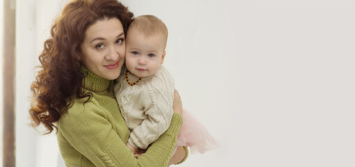 Fototapeta na wymiar A young mother in a sweater hugs the baby and looks at the camera. Portrait of a woman with a baby in her arms on a gray background near the window. The concept of care, love, family. Copy space