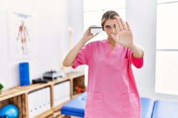 Young physiotherapist woman working at pain recovery clinic holding credit card with open hand doing stop sign with serious and confident expression, defense gesture