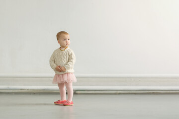 A small baby in a sweater and a fluffy skirt stands in a large empty hall against the background of a gray wall and looks to the side. Horizontal format. Copy space